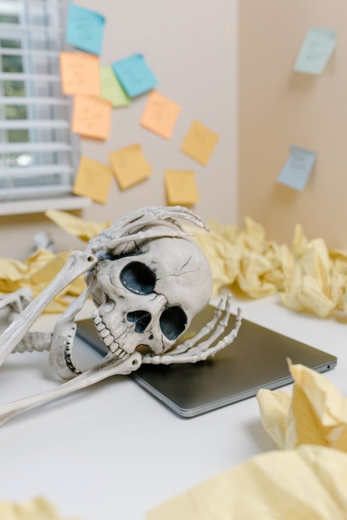 A humorous skeleton model with a laptop amid crumpled papers and sticky notes in a cluttered office setting.