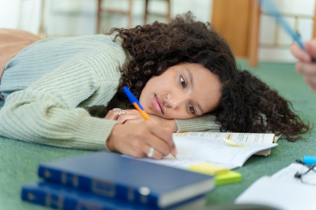 Focused woman lying on the floor writing notes, surrounded by books in a cozy indoor setting.